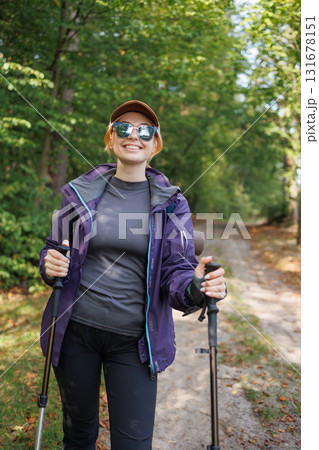 Smiling woman hiker with trekking poles walking in forest trail Smiling woman hiker with trekking poles walking in forest trail 131678151