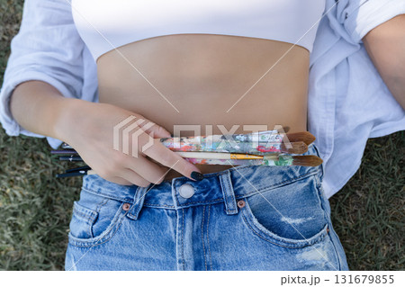 Closeup of a woman's hand tucking colourful used paintbrushes into blue jeans waistband, creative lifestyle detail with texture and casual fashion vibe, macro crop. Closeup of a woman's hand tucking colourful used paintbrushes into blue jeans waistband, creative lifestyle detail with texture and casual fashion vibe, macro crop. 131679855