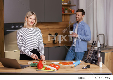 Beautiful young couple is talking and smiling while cooking healthy food in kitchen at home. Man is drinking cup of coffee and talking with woman. Beautiful young couple is talking and smiling while cooking healthy food in kitchen at home. Man is drinking cup of coffee and talking with woman. 131680665