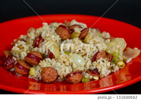 Delicious fried rice with sliced sausages and vegetables on a vibrant red plate. Delicious fried rice with sliced sausages and vegetables on a vibrant red plate. 131680682