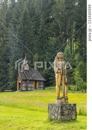 Wooden sculpture of man in meadow with rustic chapel and forest 131680894