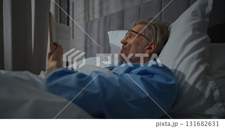 Waking Up in Morning in Bright, Cozy Bedroom, Mature Man Lies Relaxed in White Bed Reading Book Waking Up in Morning in Bright, Cozy Bedroom, Mature Man Lies Relaxed in White Bed Reading Book 131682631