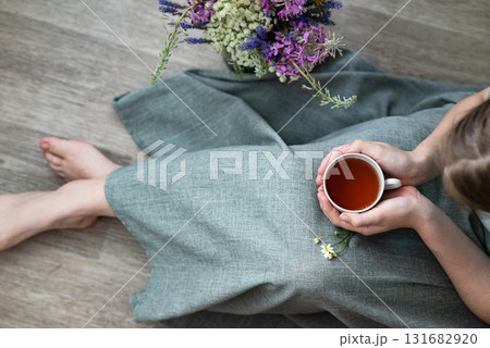 Relaxing with a cup of herbal tea while seated on the floor surrounded by fresh flowers in a cozy indoor setting Relaxing with a cup of herbal tea while seated on the floor surrounded by fresh flowers in a cozy indoor setting 131682920