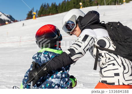 Mother teaching child to ski on sunny mountain slope, holding and guiding for balance and confidence. Warm family moment of encouragement and teamwork in bright winter landscape full of joy Mother teaching child to ski on sunny mountain slope, holding and guiding for balance and confidence. Warm family moment of encouragement and teamwork in bright winter landscape full of joy 131684188