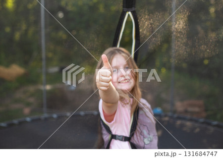 Happy excited child girl sitting on trampoline outdoor showing thumb up sign 131684747