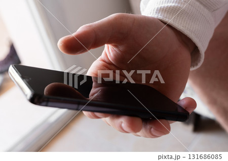 A man holds a smartphone in his hands. The phone has a sleek design and a dark screen. The background is blurred, focusing on the phone and hands. 131686085