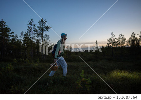 Woman walks in autumn swamp forest with basket mushrooms at dusk, lighting way with headlamp on head 131687364