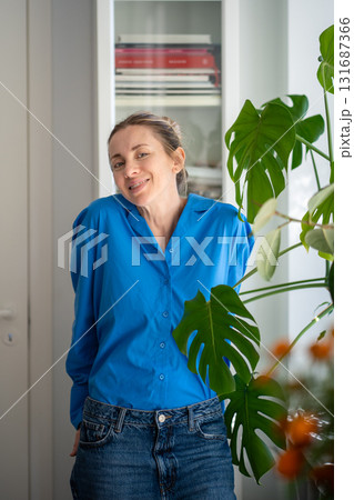 Portrait of charming smiling middle aged woman stands next to monstera leaves at cozy home Portrait of charming smiling middle aged woman stands next to monstera leaves at cozy home 131687366