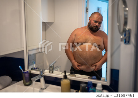 Upset man has extra weight looking at mirror touching big abdomen in bathroom at home.  131687461