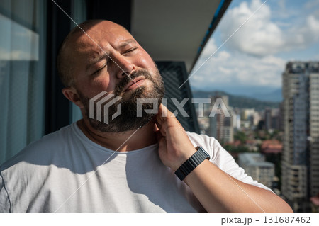 Bearded man in summer sun on balcony. Guy closed eyes in displeasure discomfort holding on to neck, 131687462