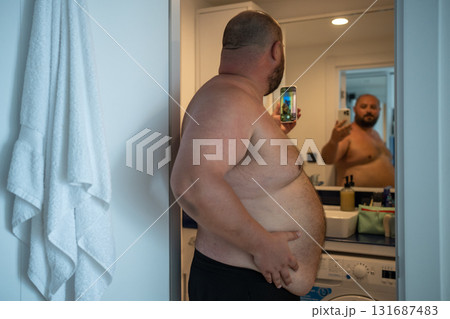 Overweight man with naked torso taking selfie on smartphone looking at mirror in bathroom at home. Overweight man with naked torso taking selfie on smartphone looking at mirror in bathroom at home. 131687483