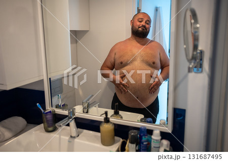 Fat man looking at mirror smiling touching big abdomen in bathroom at home.  131687495