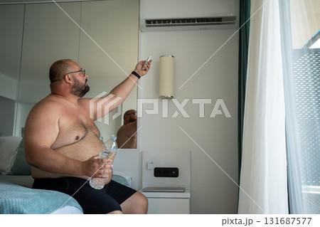 Overweight man switching on air conditioner in hotel room for cooling in hot weather on vacation. Overweight man switching on air conditioner in hotel room for cooling in hot weather on vacation. 131687577