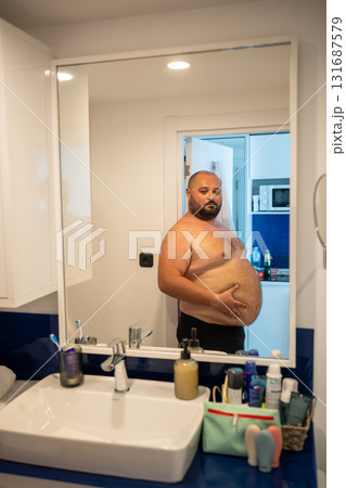 Overweight smiling man with naked torso suffering from extra weight looks at mirror touches abdomen. 131687579
