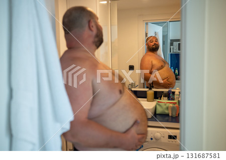 Overweight smiling man with naked torso suffering from extra weight looks at mirror touches abdomen. Overweight smiling man with naked torso suffering from extra weight looks at mirror touches abdomen. 131687581