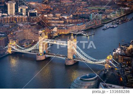 A stunning aerial view captures Tower Bridge illuminated at dusk, with the River Thames flowing beneath and city lights reflecting in the water, highlighting London's vibrant evening atmosphere. 131688286