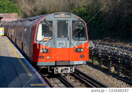 A red and grey train comes into view at a London Underground station. It is a clear day, and trees provide a green backdrop as passengers wait on the platform. A red and grey train comes into view at a London Underground station. It is a clear day, and trees provide a green backdrop as passengers wait on the platform. 131688287