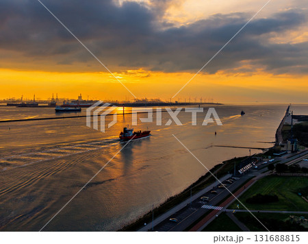 Aerial sunset view of industrial seascape ship leaves wake across golden water as cranes and port structures silhouette against dramatic clouds and warm horizon light. Aerial sunset view of industrial seascape ship leaves wake across golden water as cranes and port structures silhouette against dramatic clouds and warm horizon light. 131688815