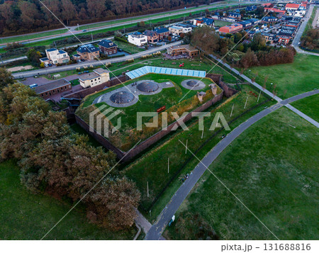 Aerial view of Fort bij Veldhuis grass covered bunkers within brick walls, adjacent to glass roofed museum, surrounded by fields, homes, and highway, blending heritage with modern infrastructure. 131688816