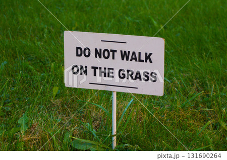 "Do not walk on the grass" sign mounted on a white rectangular board. The sign is set against a background of green grass. "Do not walk on the grass" sign mounted on a white rectangular board. The sign is set against a background of green grass. 131690264