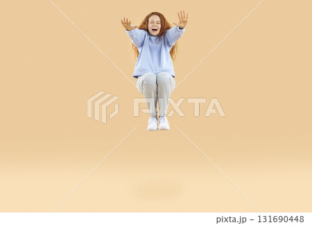 Beautiful happy red-haired young woman in light blue sweatshirt and gray jeans, squats in jump and stretches her arms forward. 131690448