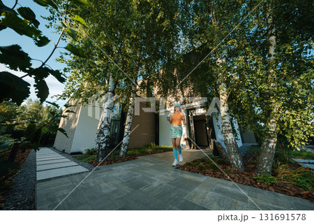 Young person walks towards modern house surrounded by greenery, showcasing a bright exterior and well-maintained garden in the afternoon light 131691578