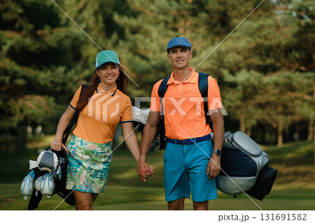 Couple enjoying a sunny day on the golf course, showcasing vibrant outfits while holding hands and carrying golf bags 131691582