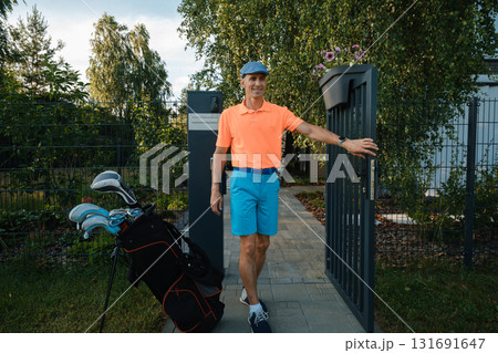 Man in bright attire stands at garden gate with golf clubs ready for a game in a residential area during late afternoon 131691647
