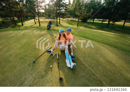 Couple enjoys a relaxing moment on the golf course in the late afternoon sun surrounded by lush greenery and golf equipment 131692160