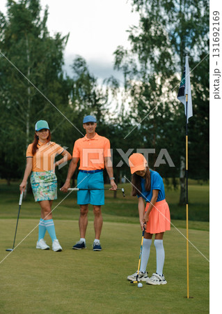 Family enjoys a fun day of golfing on a sunny afternoon at the course, with a youngster preparing to putt as parents watch and cheer 131692169