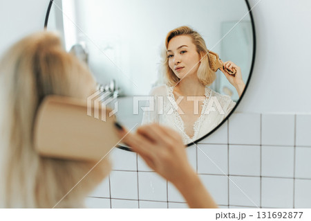 Back view of beautiful young woman in white undershirt combing her hair and smiling while looking into the mirror in bathroom 131692877
