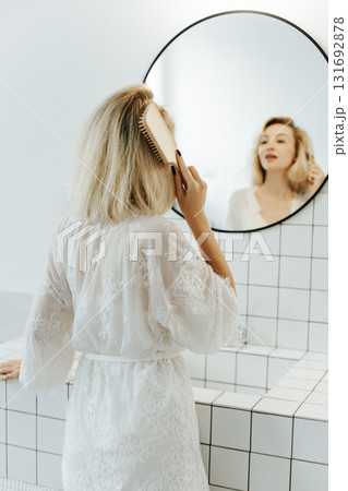 Back view of beautiful young woman in white undershirt combing her hair and smiling while looking into the mirror in bathroom 131692878