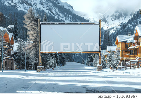 Blank billboard stands on snow-covered street between wooden cabins with snowy mountains in background. Concept of advertising, winter, nature. For winter holiday promotion 131693967
