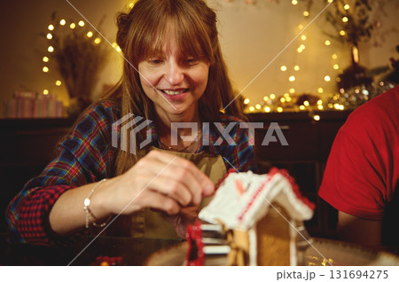 Woman Decorating A Gingerbread House With Warm Christmas Lights And Festive Decor, Sharing A Cozy Holiday Moment At The Table Woman Decorating A Gingerbread House With Warm Christmas Lights And Festive Decor, Sharing A Cozy Holiday Moment At The Table 131694275