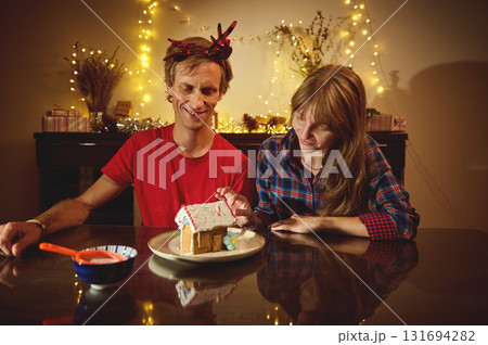 Happy Couple Celebrates Christmas Evening, Decorating Gingerbread House With Warm Lights and Cozy Festive Decor 131694282