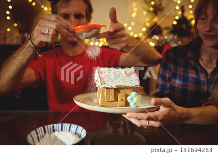 Family And Friends Decorate A Gingerbread House For Christmas, Dusting Sugar On The Roof As Warm Lights Create A Cozy, Festive Moment 131694283