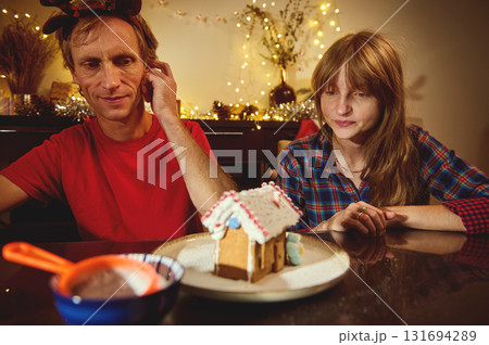 Couple At Home Celebrating Christmas With Gingerbread House Craft, Soft Fairy Lights In The Background, Cozy Holiday Vibe As They Sit At The Table And Share A Sweet Moment 131694289