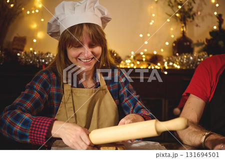 Joyful Baker in Chef Hat and Apron Rolling Dough During Cozy Christmas Kitchen Scene Joyful Baker in Chef Hat and Apron Rolling Dough During Cozy Christmas Kitchen Scene 131694501