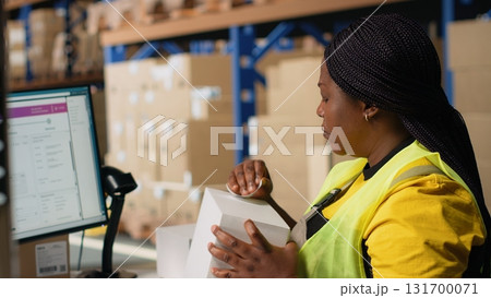 African american woman printing and applying airway bill tags on boxes, wearing hi vis vest in a warehouse near racks. Managing barcode labels with tracking info for order fulfillment. Camera B. 131700071