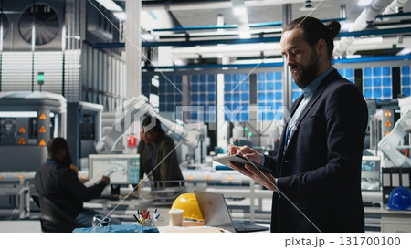Industrial male expert in suit inspecting solar power equipment, demonstrating efficiency in quality control and production line. Working on solar panels manufacturing. Camera A. 131700100