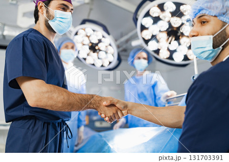 Two surgeons shake hands in an operating room, likely before or after a procedure, with medical staff and equipment in the background. 131703391
