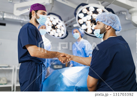 Two surgeons shake hands in an operating room, with other medical staff and surgical equipment in the background. 131703392