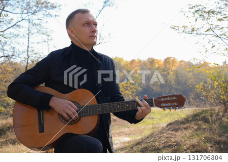 Blonde man playing string guitar outdoors in autumn forest. Concept of sound therapy, mental health and wellness rituals. Calmness tranquility audio-sensory practices. Aura farming energy Blonde man playing string guitar outdoors in autumn forest. Concept of sound therapy, mental health and wellness rituals. Calmness tranquility audio-sensory practices. Aura farming energy 131706804