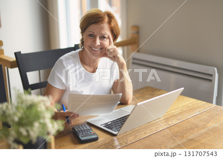 happy senior woman calculating family budget, sitting at kitchen counter with laptop, papers, 131707543