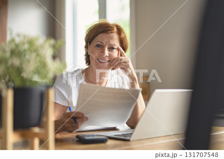 happy senior woman calculating family budget, sitting at kitchen counter with laptop, papers, 131707548