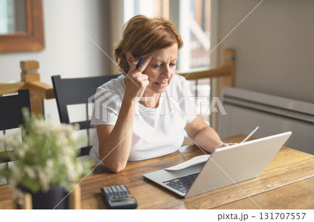 Sad frustrated senior woman pensioner having depressed look, holding hand on her face, calculating family budget, sitting at kitchen counter with laptop, papers, coffee, calculator and cell phone 131707557