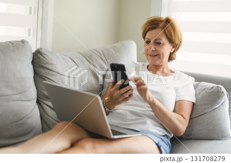 senior woman relaxing at home on the sofa reading her text messages on her mobile phone with a quiet smile 131708279