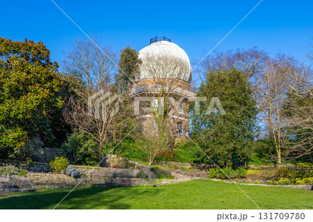 Visitors enjoy the scenic beauty of the Old Royal Observatory in Greenwich, surrounded by lush green gardens and trees, under a clear blue sky on a sunny day. 131709780