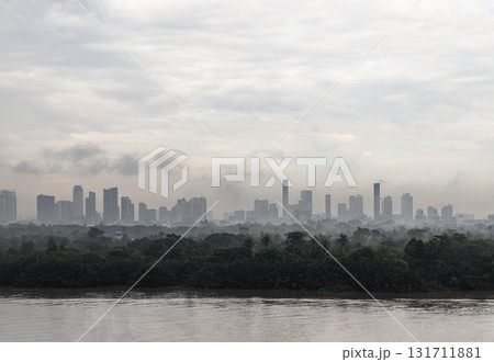 a cityscape visible through a haze after the rain with a body of water on the Chao Phraya River and a forested area of Bang Krachao in the foreground. a cityscape visible through a haze after the rain with a body of water on the Chao Phraya River and a forested area of Bang Krachao in the foreground. 131711881