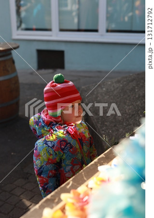 Little girl in colorful winter clothes and striped hat gazes at festive display at Christmas market, lit by sunlight 131712792